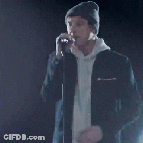A Man In A Beanie Is Singing Into A Microphone With The Words Our Time Written On It . GIF
