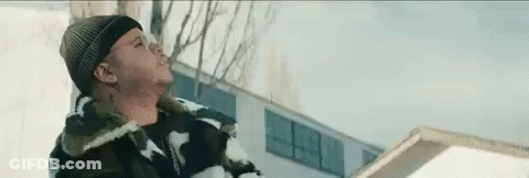 A Man In A Beanie Is Standing In The Snow Near A Building . GIF