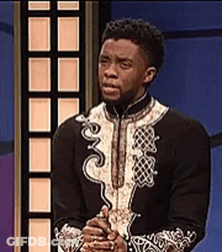 A Man In A Black And White Outfit Is Standing In Front Of A Blue Wall With His Hands Folded . GIF