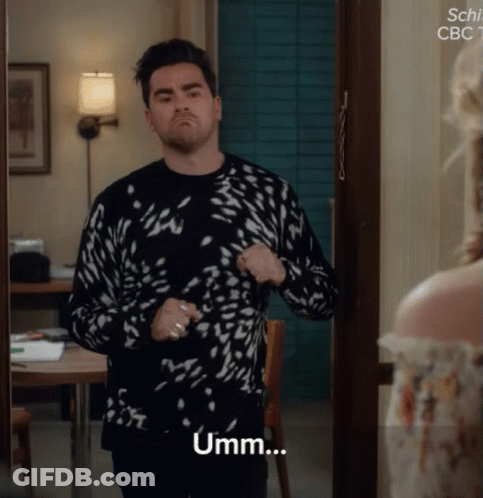 A Man In A Black And White Sweater Stands In Front Of A Door And Says Umm GIF