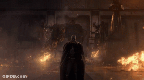A Man In A Black Cape Is Standing In Front Of A Burning Building GIF