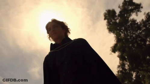 A Man In A Black Cape Stands In Front Of A Tree With The Sun Shining Through It GIF