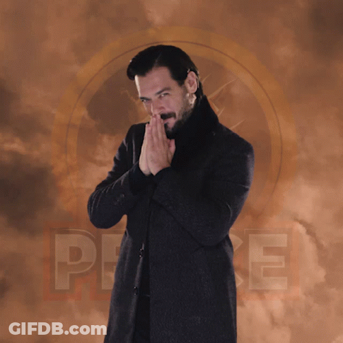 A Man In A Black Coat And Black Turtleneck Stands In Front Of A Cloudy Background With The Letter P On It GIF