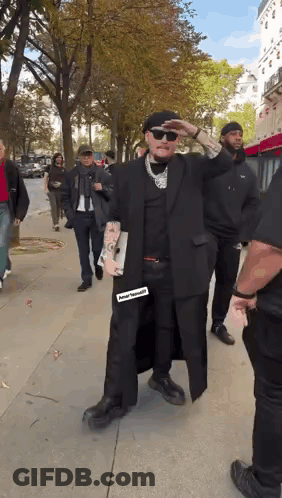 A Man In A Black Coat And Hat Is Walking Down A Sidewalk With A Group Of People . GIF