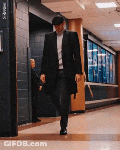 A Man In A Black Coat And Hat Walks Down A Hallway With A Sign On The Wall That Says ' Nfl ' GIF