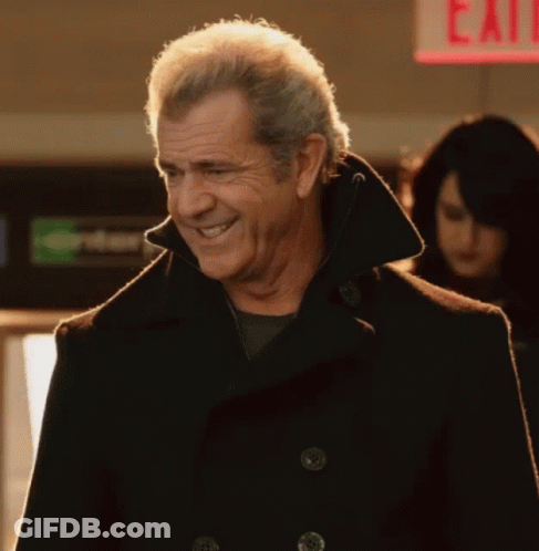 A Man In A Black Coat Is Smiling In Front Of A Red Exit Sign GIF