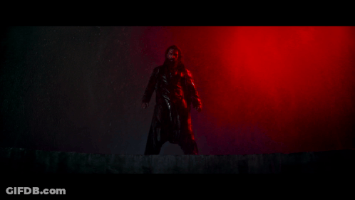 A Man In A Black Coat Is Standing In A Dark Room With Red Lights Behind Him . GIF