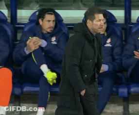A Man In A Black Coat Is Walking In Front Of A Row Of Players GIF