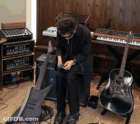 A Man In A Black Coat Sits In Front Of A Keyboard And Guitar GIF