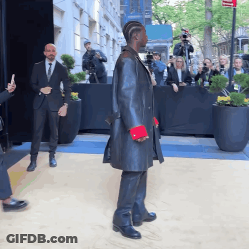 A Man In A Black Coat With Red Cuffs Stands In Front Of A Crowd GIF