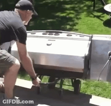 A Man In A Black Hat Is Pushing A Cart With A Grill In The Background GIF