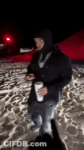 A Man In A Black Hooded Jacket Is Standing On A Beach Holding A Cup . GIF