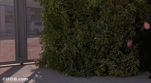 A Man In A Black Hooded Jacket Stands In Front Of A Fence GIF