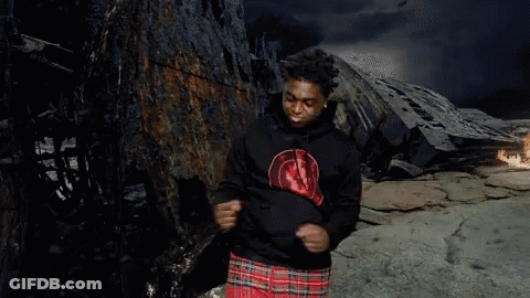 A Man In A Black Hoodie And Plaid Shorts Is Standing In Front Of A Burned Out Building . GIF