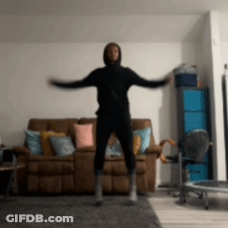 A Man In A Black Hoodie Is Jumping In Front Of A Couch GIF