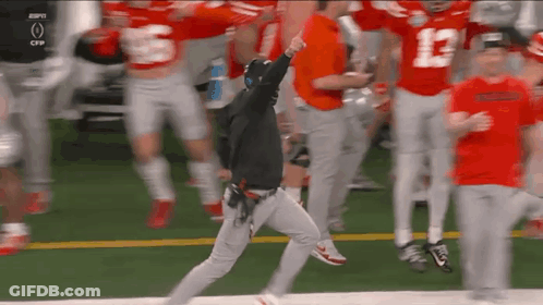 A Man In A Black Hoodie Is Running On A Football Field Surrounded By Football Players . GIF