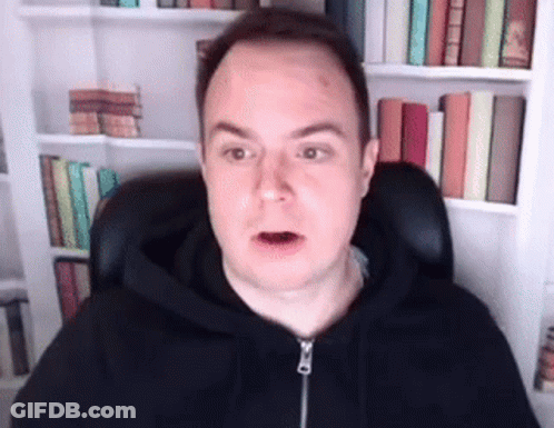 A Man In A Black Hoodie Is Sitting In Front Of A Bookshelf And Making A Funny Face . GIF