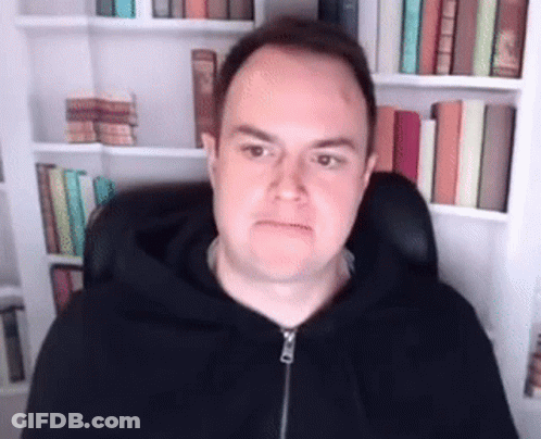 A Man In A Black Hoodie Is Sitting In Front Of A Bookshelf With Books . GIF