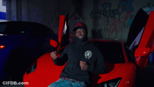 A Man In A Black Hoodie Is Sitting In Front Of A Red Sports Car GIF