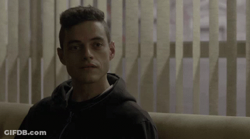 A Man In A Black Hoodie Is Sitting On A Couch In Front Of Vertical Blinds GIF