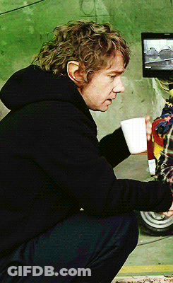 A Man In A Black Hoodie Is Squatting Down Holding A Cup Of Coffee GIF