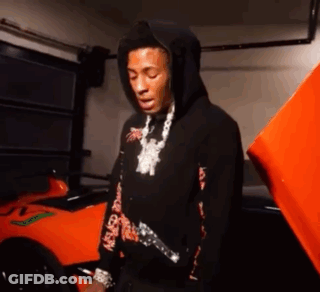 A Man In A Black Hoodie Is Standing In Front Of An Orange Car In A Garage . GIF