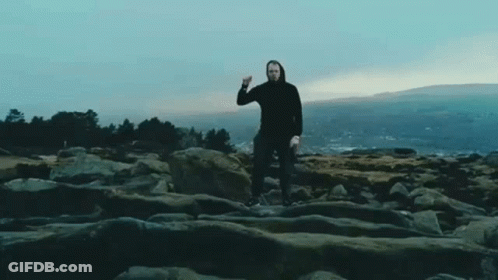 A Man In A Black Hoodie Is Standing On A Rocky Cliff With A Mountain In The Background . GIF