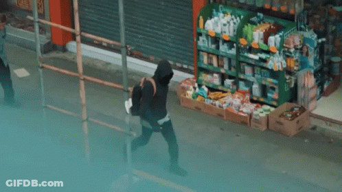 A Man In A Black Hoodie Is Walking Down A Sidewalk In Front Of A Store . GIF
