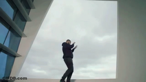 A Man In A Black Hoodie Stands In Front Of A Large Window GIF