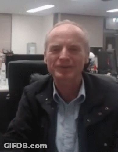 A Man In A Black Jacket And Blue Shirt Is Sitting At A Desk In An Office GIF