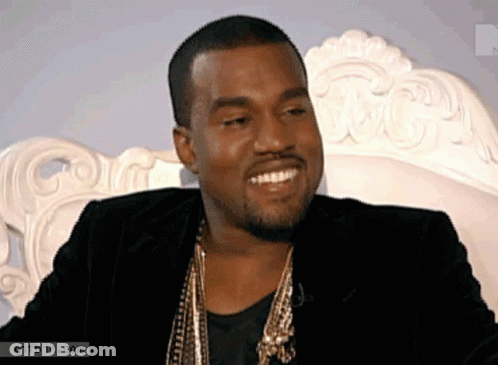 A Man In A Black Jacket And Gold Necklace Smiles While Sitting On A White Couch GIF