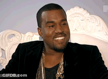 A Man In A Black Jacket And Gold Necklace Smiles While Sitting On A White Couch GIF