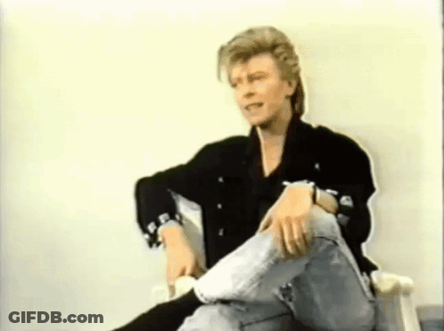 A Man In A Black Jacket And Jeans Is Sitting In A Chair With His Legs Crossed . GIF