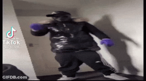 A Man In A Black Jacket And Purple Gloves Is Standing In A Room . GIF