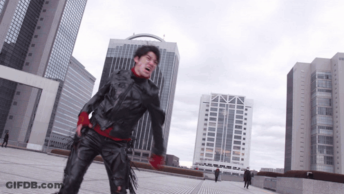 A Man In A Black Jacket And Red Gloves Stands In Front Of A City Skyline GIF