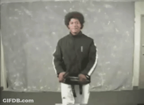 A Man In A Black Jacket And White Pants Is Standing In Front Of A White Wall Holding A Black Belt . GIF