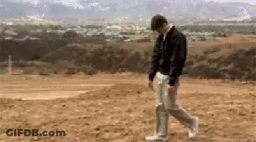 A Man In A Black Jacket And White Pants Is Walking On A Dirt Field . GIF