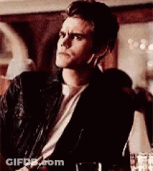 A Man In A Black Jacket And White Shirt Is Sitting In A Room . GIF