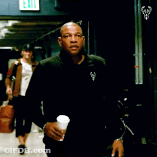 A Man In A Black Jacket Holds A Cup Of Coffee In Front Of An Exit Sign GIF