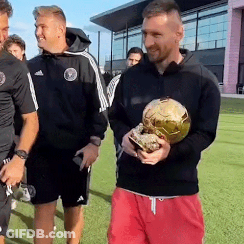 A Man In A Black Jacket Is Holding A Gold Soccer Ball While Standing On A Field . GIF