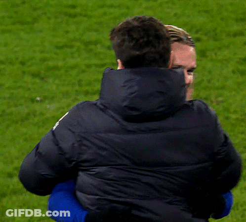 A Man In A Black Jacket Is Hugging Another Man In A Blue Shirt GIF