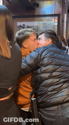 A Man In A Black Jacket Is Kissing Another Man In An Orange Shirt GIF