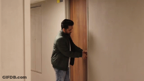 A Man In A Black Jacket Is Opening A Door In A Hallway GIF