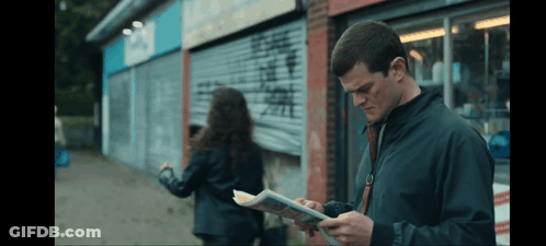 A Man In A Black Jacket Is Reading A Newspaper While A Woman Looks On GIF