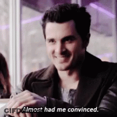 A Man In A Black Jacket Is Sitting At A Table And Smiling While Saying `` Almost Had Me Convinced . '' GIF