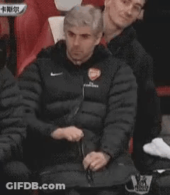 A Man In A Black Jacket Is Sitting In A Chair With His Hands Folded . GIF