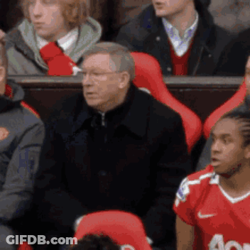 A Man In A Black Jacket Is Sitting In A Stadium With Other People GIF