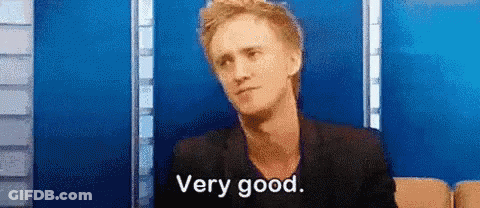 A Man In A Black Jacket Is Sitting In Front Of A Blue Wall And Saying `` Very Good '' . GIF