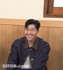 A Man In A Black Jacket Is Smiling In Front Of A Wooden Wall . GIF