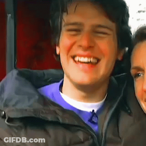 A Man In A Black Jacket Is Smiling While Standing Next To A Woman In A Purple Shirt . GIF